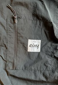 Pungă din țesătură neagră cu fermoar, având o etichetă albă rectangulară pe care este imprimată marca "einy" și "unbored onboard" cu litere negre.