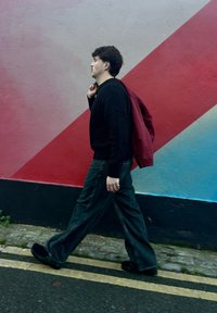 Un uomo con maglione nero e jeans cammina davanti a un muro colorato, portando una giacca rossa sulla spalla, su una strada con linee gialle doppie.