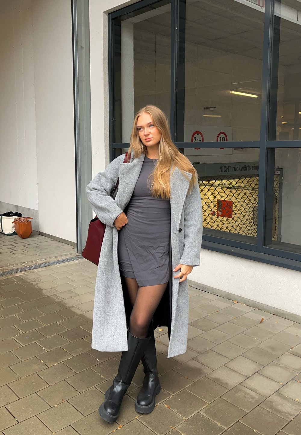 Cappotto di lana grigio sopra un vestito grigio, stivali neri con tacco alto fino al ginocchio e borsa bordeaux. Texture lisce e design strutturato.