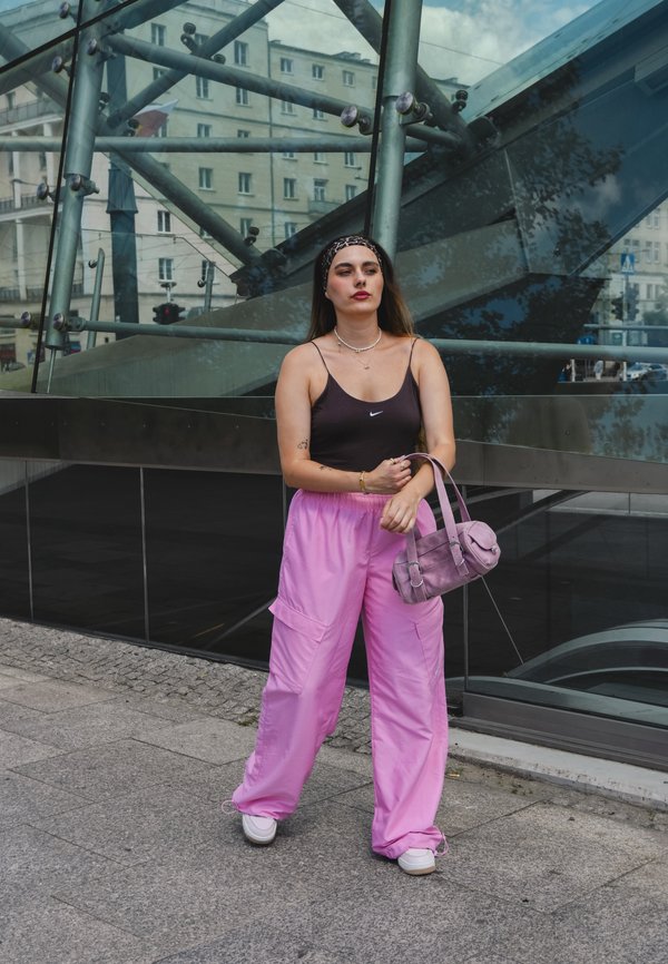 Top nero senza maniche, pantaloni larghi rosa, sneakers bianche e una borsa lavanda. Capelli lunghi castani, gioielli semplici e una fascia per capelli.