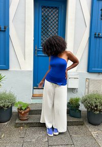 Bluo top a spalla unica con texture arricciata, pantaloni bianchi a gamba larga e scarpe da ginnastica blu. Circondata da piante in vaso e da una porta blu.