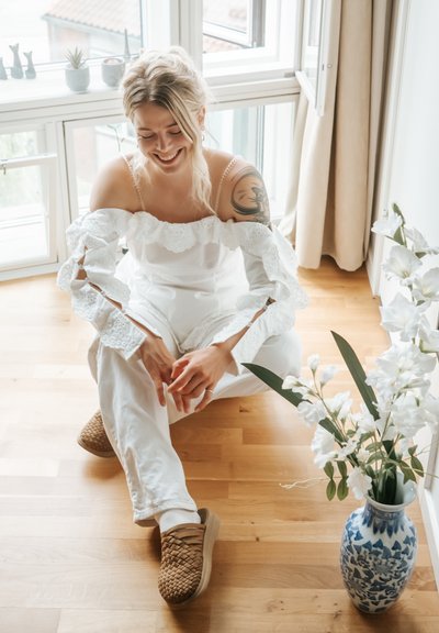 Blusa blanca sin mangas con encaje en los bordes y mangas largas, combinada con pantalones blancos y zapatos cómodos de tela marrón. Jarrón azul y blanco con flores.