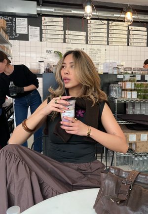 Top sin mangas negro y falda marrón, sosteniendo una bebida con un detalle de flor morada. Bolsa de cuero marrón sobre la mesa. Fondo de azulejos blancos.