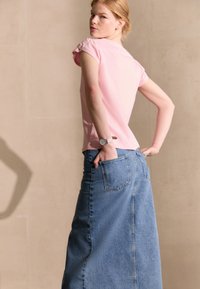 Donna con capelli rossi che indossa una camicia rosa a maniche corte e una lunga gonna di jeans blu, con le mani nelle tasche posteriori, mentre guarda oltre la spalla.