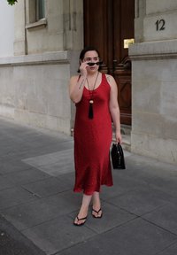 Red sleeveless dress with a textured surface, worn with black flip-flops. Holding a small black handbag, standing on a paved sidewalk.