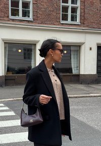 Cappotto nero su una blusa beige con bottoni, in mano una borsa in pelle bordeaux scuro. Ambiente urbano con un edificio in mattoni sullo sfondo.