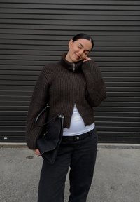 Brun ribbestrikket genser med høy krage og glidelås. Kombinert med svarte vide jeans og en svart veske. Grå bakgrunn.