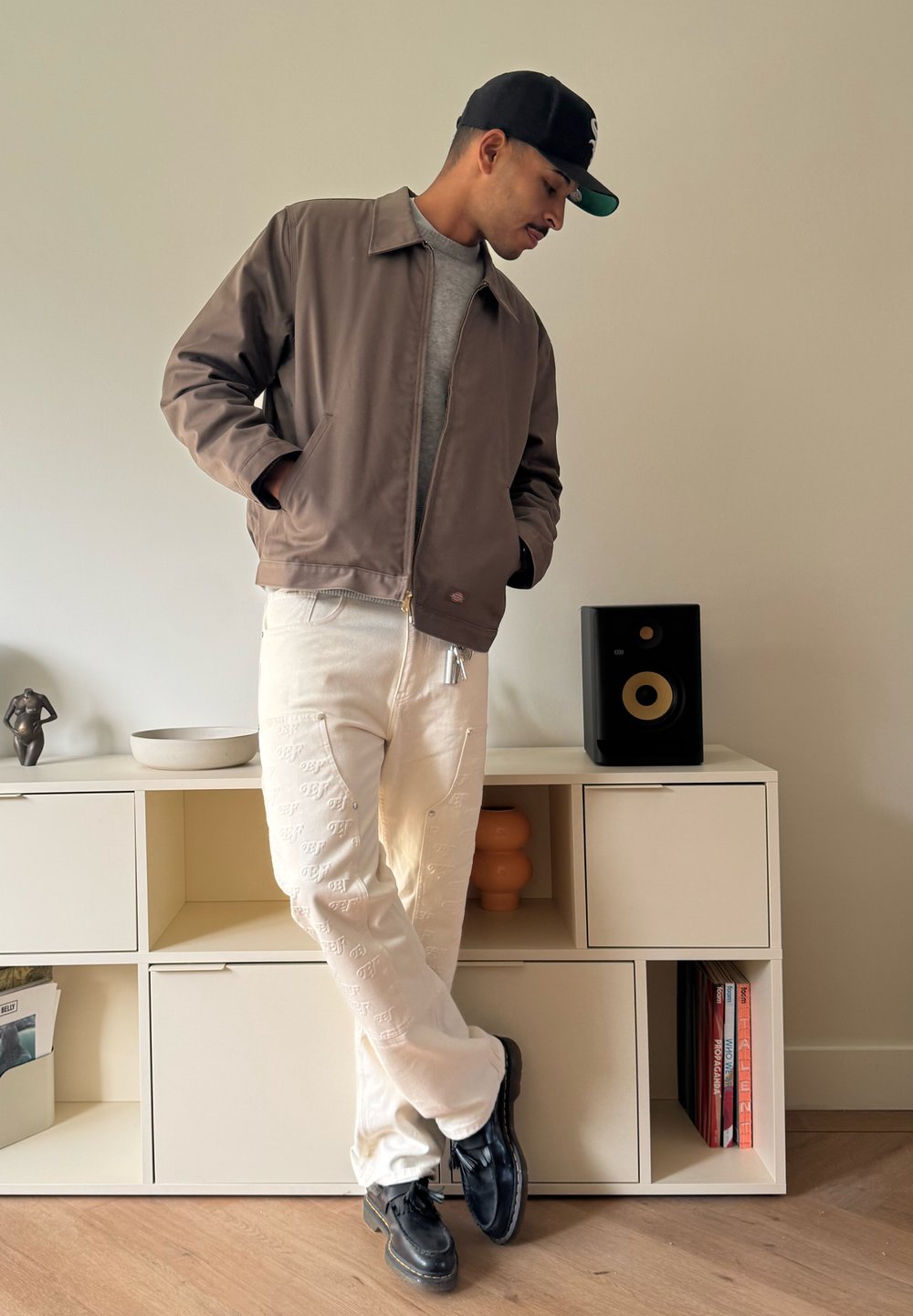 Ruskea vetoketjullinen takki, harmaa t-paita, valkoiset housut kohokuvioiduilla kuvioilla, mustat kengät; seisoo vaalean seinän vieressä modernien huonekalujen lähellä.