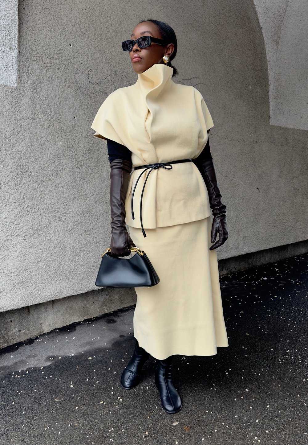 Femme en cape beige à col montant et jupe, ceinture noire, longs gants noirs, bottes noires, lunettes de soleil et petit sac à main noir, debout sur le trottoir.