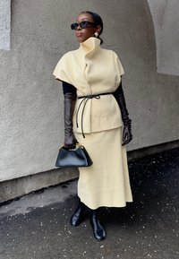 Femme en cape beige à col montant et jupe, ceinture noire, longs gants noirs, bottes noires, lunettes de soleil et petit sac à main noir, debout sur le trottoir.