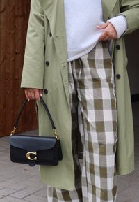 Μαύρη δερμάτινη τσάντα με χρυσό υλικό, ορθογώνιο σχήμα και κοντό χερούλι. Το μοντέλο φοράει ένα ανοιχτό πράσινο παλτό και καρό παντελόνι.