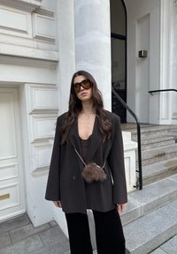 Oversized donkerblazer met krijtstrepen en brede mouwen, gelaagd over een bruin topje, gecombineerd met zwarte broek. Heeft een tas met bontaccenten aan een ketting.