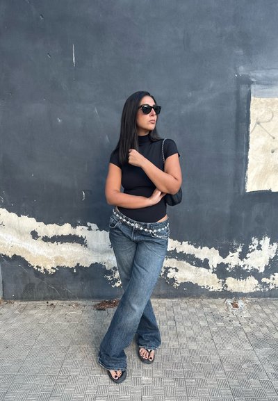 Top negro ajustado, jeans azules claros desgastados con cinturón decorativo de cadena, sandalias negras y gafas de sol, apoyado contra una pared gris texturizada.