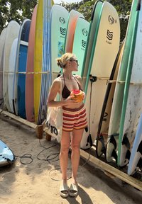Vrouw in gestreepte korte broek en bikini top die een kokosnoot vasthoudt, blootsvoets staand bij een rek met kleurrijke surfplanken op zandgrond.