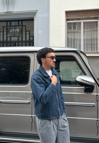 Denim jacket in dark blue with zip closure, grey wide-leg trousers, and sunglasses, standing next to a silver SUV against a backdrop of a building.