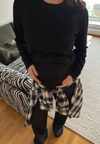 Fekete, hosszú ujjú pulóver fekete nadrág fölött, derékon átkötött szürke és fekete kockás inggel, kezek a hason.
