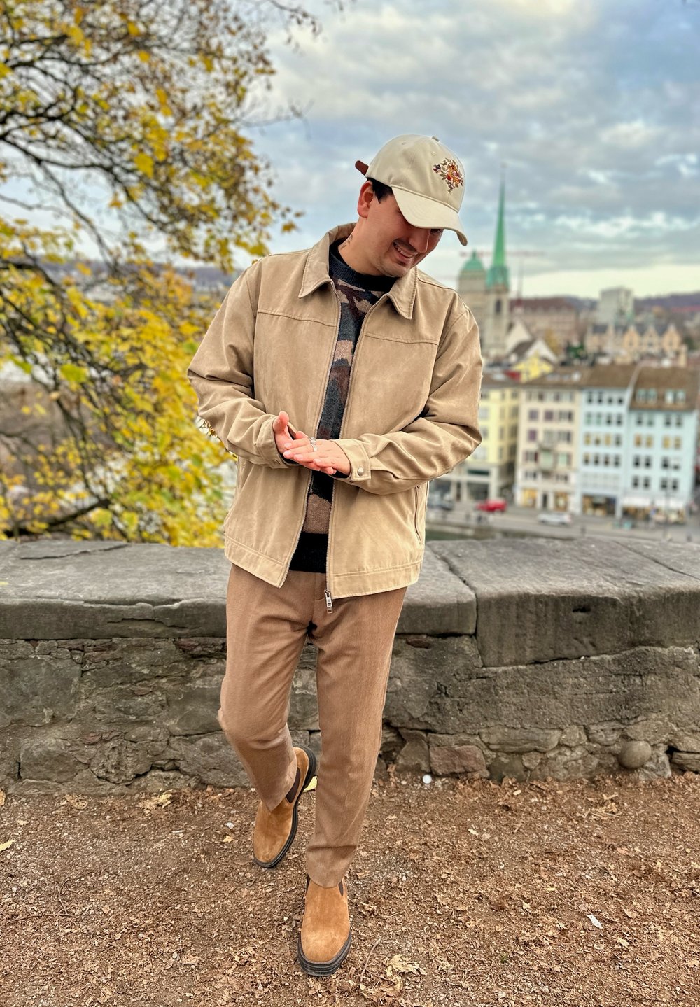 Beige Wildlederjacke über einem gemusterten Pullover, braune Hose und hellbraune Wildlederschuhe. Accessoires beinhalten eine hellfarbige Mütze mit grafischem Design.