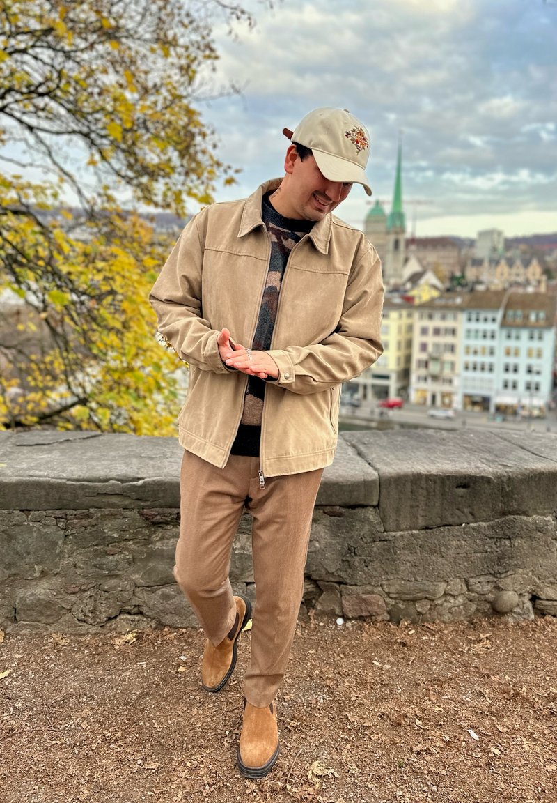 Beige suede jacket over a patterned jumper, brown trousers, and tan suede shoes. Accessories include a light-coloured cap with a graphic design.