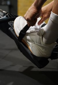 Χέρια που ασφαλίζουν μαύρο λουρί πάνω σε λευκό αθλητικό παπούτσι στο πεντάλ μηχανής κωπηλασίας με λευκές κάλτσες σε εσωτερικό γυμναστήριο.