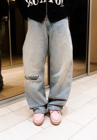 Άτομο που φορά ανοικτό μπλε σκισμένα τζιν και ροζ παπούτσια, στέκεται σε πλακόστρωτο πάτωμα με καθρεφτισμένη ντουλάπα στο φόντο.