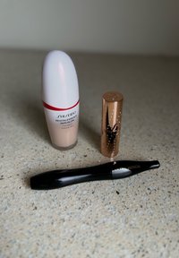 Fond de teint Shiseido dans un flacon rond blanc avec des accents rouges, un étui à rouge à lèvres en or rose avec un détail ressemblant à des diamants, et un tube de mascara noir.