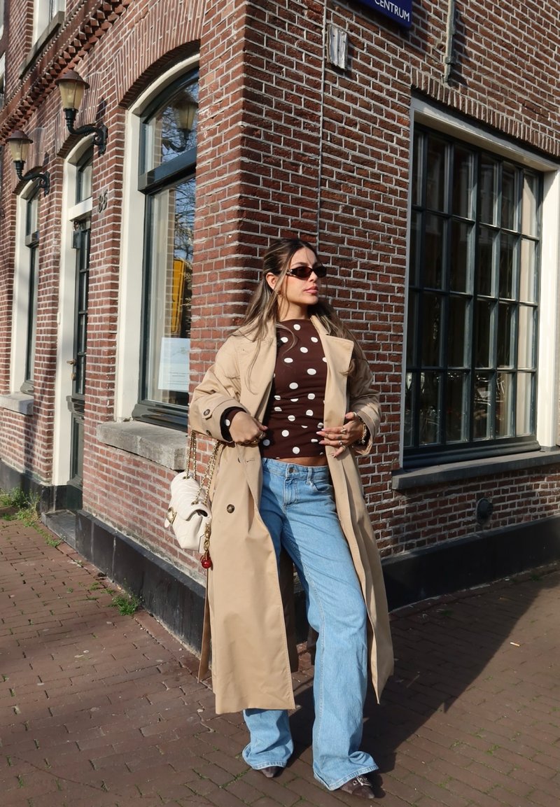 Frau trägt beigen Trenchcoat, braunes gepunktetes Oberteil, hellblaue Jeans und Sonnenbrille, steht auf einem gepflasterten Gehweg neben einem Ziegelgebäude.