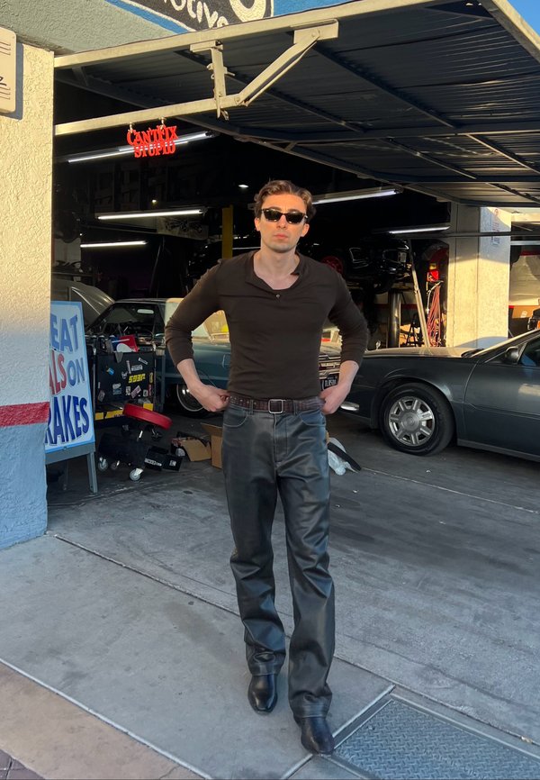 Black long-sleeved shirt, leather trousers, and sunglasses. Standing in a garage with cars and a bright sign in the background.