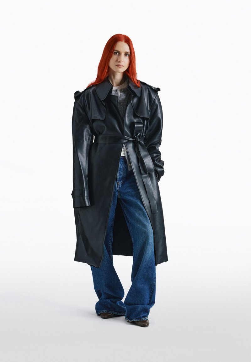Frau mit roten Haaren, die einen langen schwarzen Leder-Trenchcoat, ein graues Oberteil und weite blaue Jeans trägt, steht vor einem weißen Hintergrund.