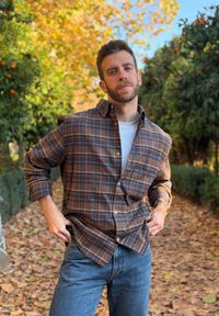 Jeune homme aux cheveux courts et à la barbe, portant une chemise à carreaux marron et un jean bleu, debout sur un sentier couvert de feuilles bordé d'orangers en automne.