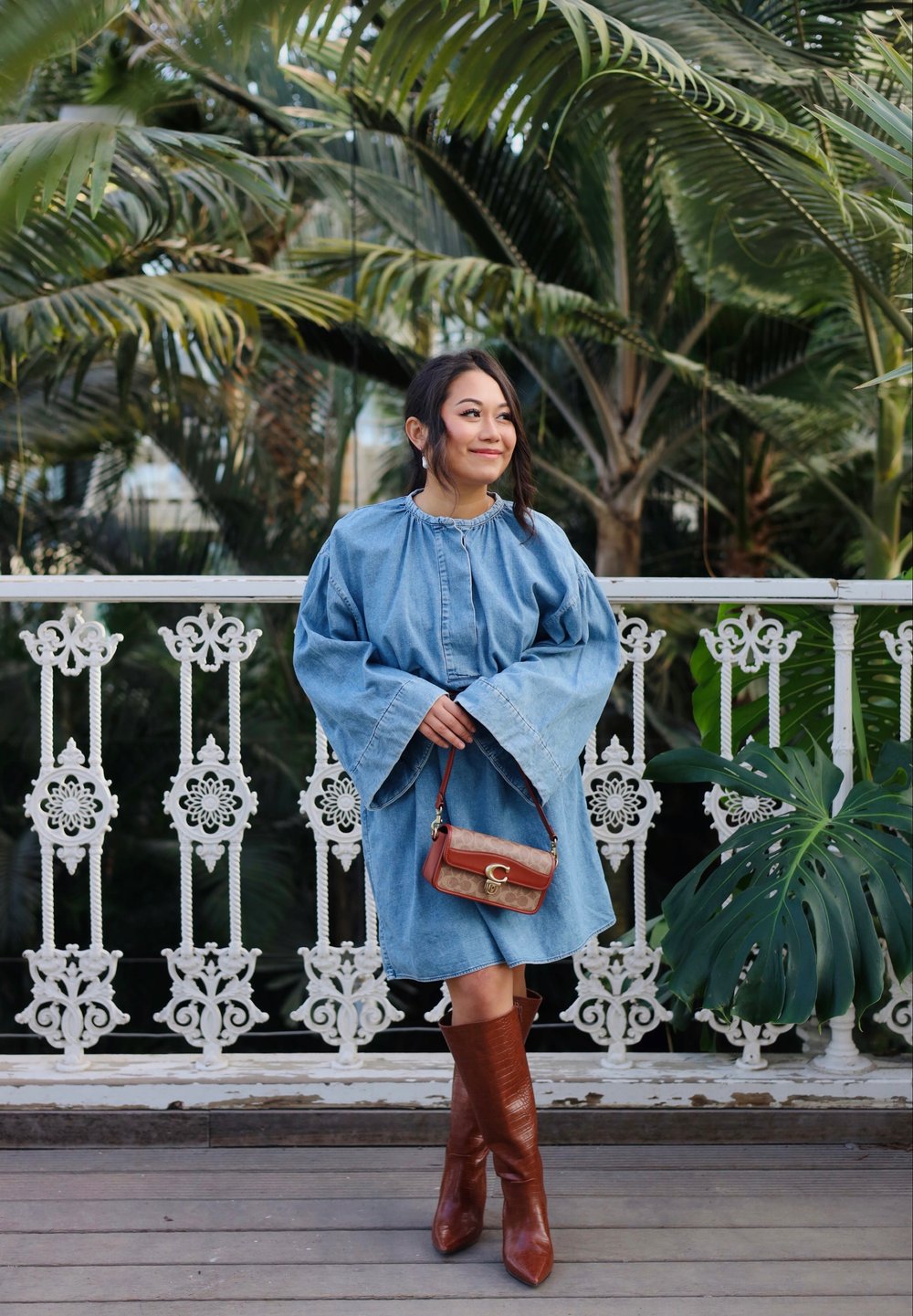 Robe en denim avec des manches évasées, couleur bleue, taille cintrée, associée à des bottes marron jusqu'au genou et un petit sac à main marron et beige avec un logo.