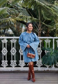 Robe en denim avec des manches évasées, couleur bleue, taille cintrée, associée à des bottes marron jusqu'au genou et un petit sac à main marron et beige avec un logo.