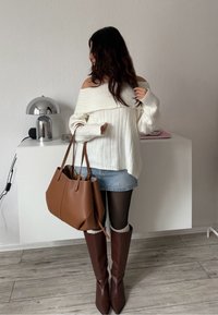 Femme portant un pull blanc épaules dénudées, jupe en denim, collants noirs, bottes marron montantes, tenant un grand sac à main marron dans une pièce minimaliste.