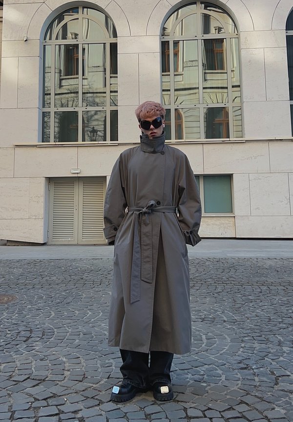 Personne aux cheveux courts roses et lunettes de soleil portant un long trench-coat gris ceinturé et des chaussures noires épaisses, debout sur une rue pavée.