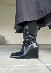 Botas de cuña negras hasta la rodilla con punta afilada, fabricadas en material brillante, que presentan un detalle de fruncido en el tobillo y un robusto tacón de cuña.
