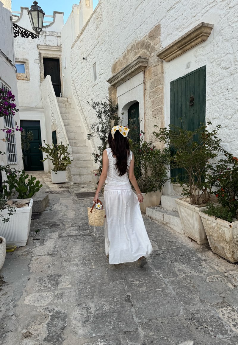Vestido de linho branco com um acabamento texturizado, combinado com uma cesta de vime. Rua de pedra ladeada por plantas em vasos e escadas que sobem.