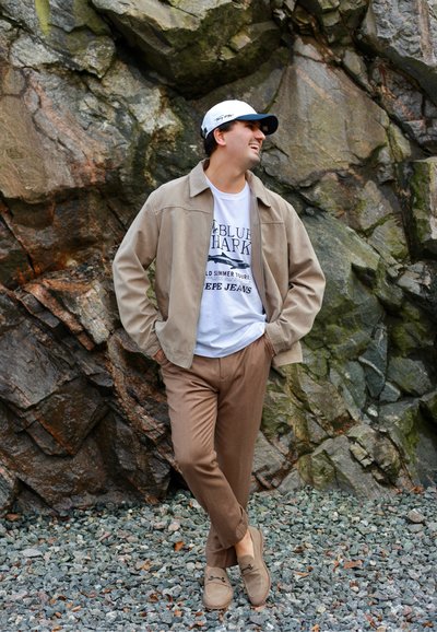 Hombre joven con chaqueta beige, pantalones marrones y gorra blanca, de pie sobre grava, sonriendo y apoyado contra una pared rocosa.