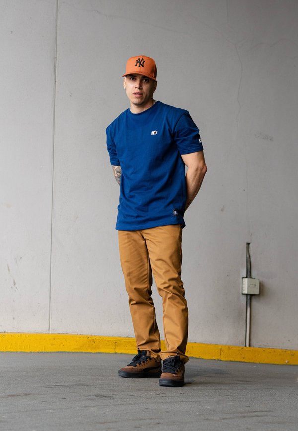Kék rövid ujjú ing, bézs slim-fit nadrág és fekete-barna sportcipő. Narancssárga baseball sapka fekete logóval. Egy szürke falnak támaszkodva áll.