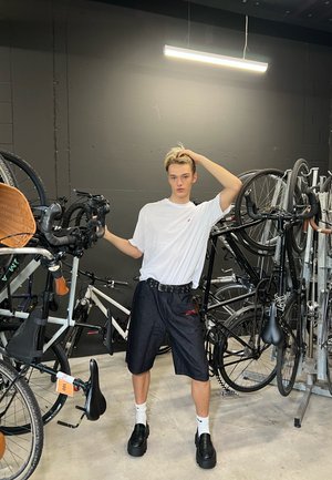 Camiseta blanca, pantalones cortos negros con logo rojo, cinturón negro. Bicicletas circundantes montadas en estantes. Suelo de concreto con poca iluminación.
