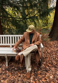 Manteau en laine brun, pull vert, pantalon beige et mocassins marron. Tenant une tasse de café, assis sur un banc blanc au milieu des feuilles d'automne.
