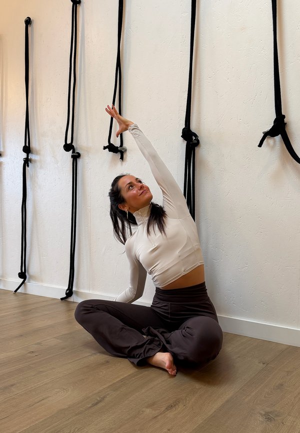 Γυναίκα με ρούχα γυμναστικής καθισμένη με τα πόδια σταυρωμένα σε ξύλινο πάτωμα, εκτελεί πλευρική διάταση με σχοινιά που κρέμονται σε λευκό τοίχο πίσω της.