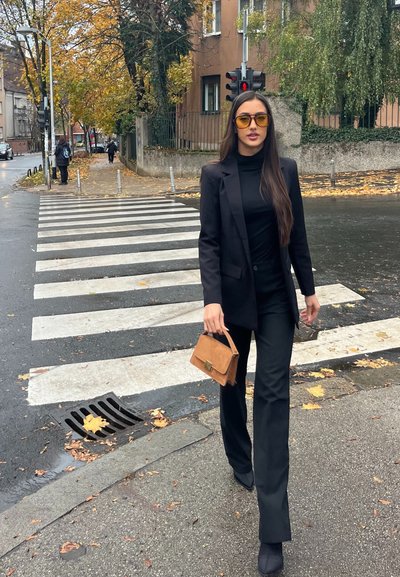 Blazer negro a medida sobre un suéter de cuello alto negro, combinado con pantalones negros. Un bolso marrón con detalle de cierre. Paso de peatones y hojas de otoño visibles.