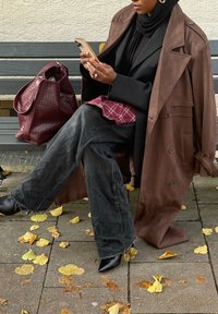 Cappotto trench in suede marrone sopra un blazer nero, indossato con una camicia a quadri e jeans larghi scuri. Porta una borsa intrecciata bordeaux e stivaletti neri.