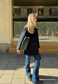 Juodas dvigubas paltas, mėlyni flared džinsai, leopardų rašto batai ir žalias popierinis pirkinių maišelis. Saulėta lauko aplinka su raštuotu langu.