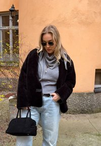 Femme portant une veste noire, une écharpe grise et un haut, un jean bleu clair, des lunettes de soleil, tenant un sac à main noir, debout à l'extérieur près d'un mur orange et d'une fenêtre.