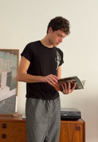 Black short-sleeve T-shirt with geometric white design, grey loose-fit trousers, holding a magazine while standing next to wooden furniture.