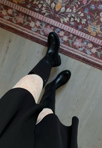 Cizme negre până la genunchi, cu finisaj neted și tălpi groase, purtate cu dresuri cu model din dantelă și o fustă neagră, pe un podea texturată.