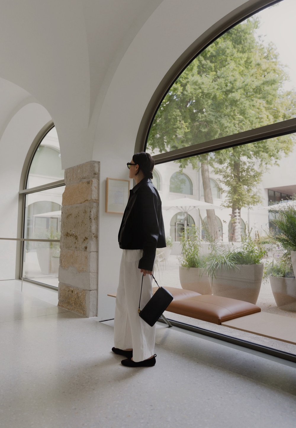 Sort cropped jakke, hvide vide bukser og sort taske. Glatte teksturer, minimalistisk design, store buede vinduer med grønne planter udenfor.