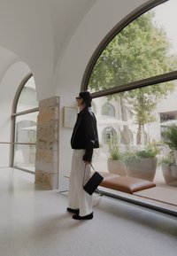 Sort cropped jakke, hvide vide bukser og sort taske. Glatte teksturer, minimalistisk design, store buede vinduer med grønne planter udenfor.