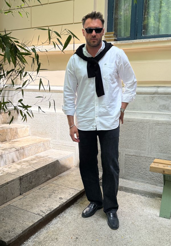 Άνδρας με μαύρα γυαλιά ηλίου, λευκό πουκάμισο με κουμπιά και μαύρο πουλόβερ περασμένο στους ώμους, μαύρο παντελόνι, στέκεται σε πετρόχτιστα σκαλοπάτια έξω.