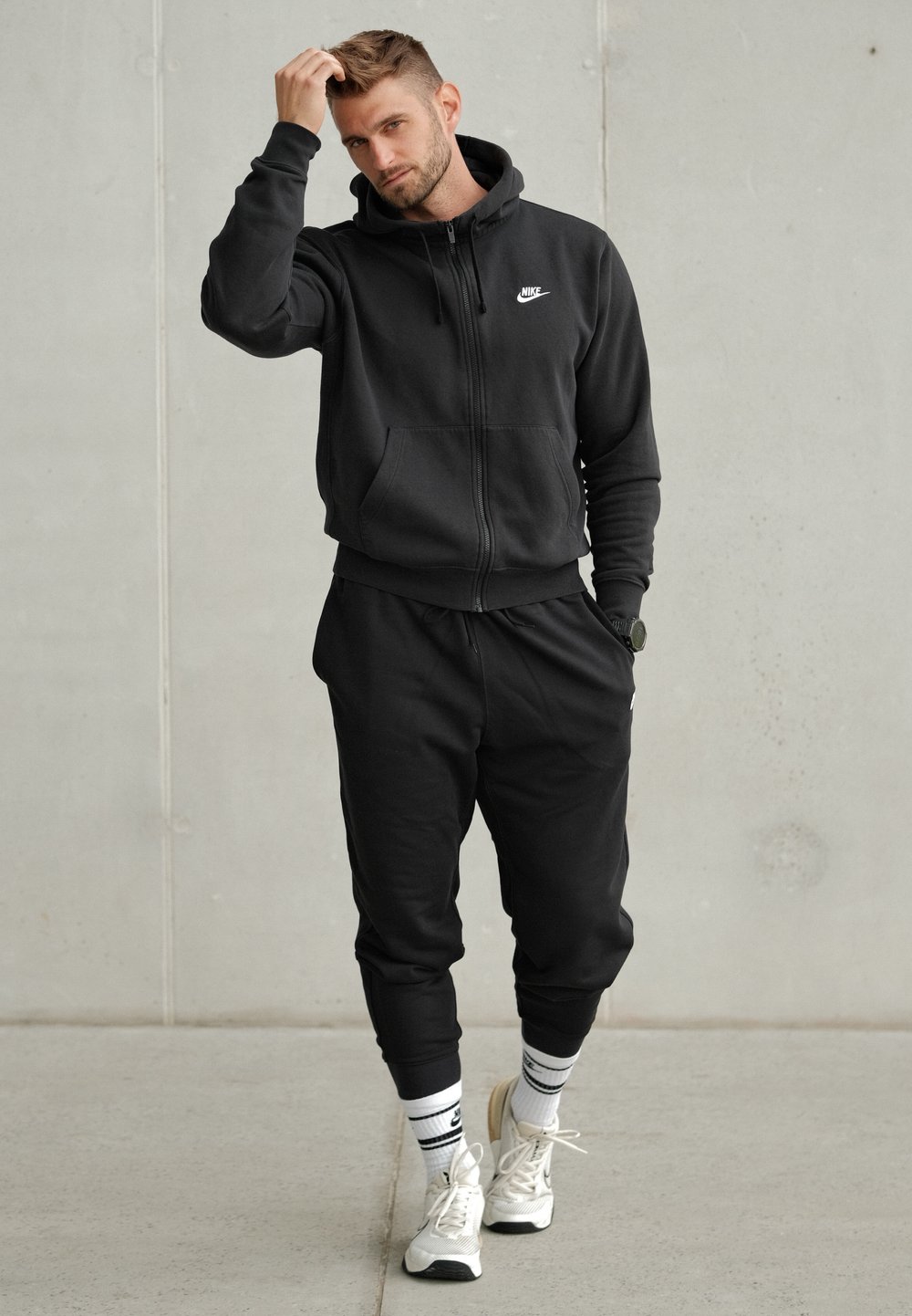 Glugă Nike neagră cu fermoar, dotată cu buzunare tip kangaroo, asortată cu pantaloni jogger. Purtați cu adidași albi și șosete cu dungi. Design minimalist.